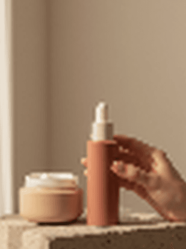 A close editorial crop of a skincare bottle and cream jar arranged in warm window light.