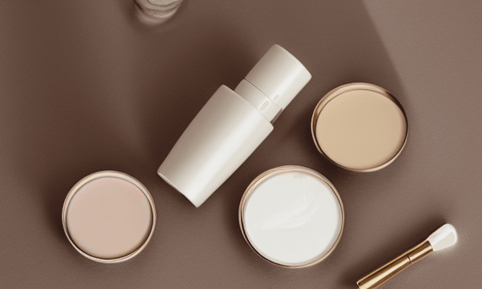 A calm editorial skincare flat lay with a serum bottle, cream jars, and a brush on a muted clay surface.
